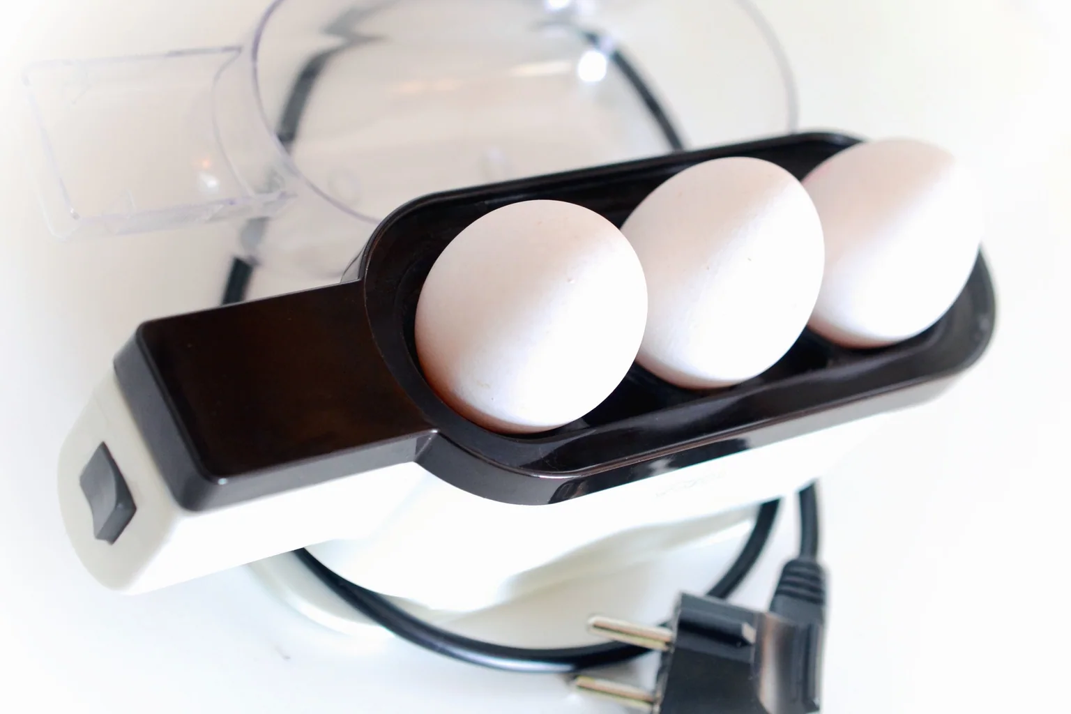 Electric egg cooker holding three eggs beside its power cord