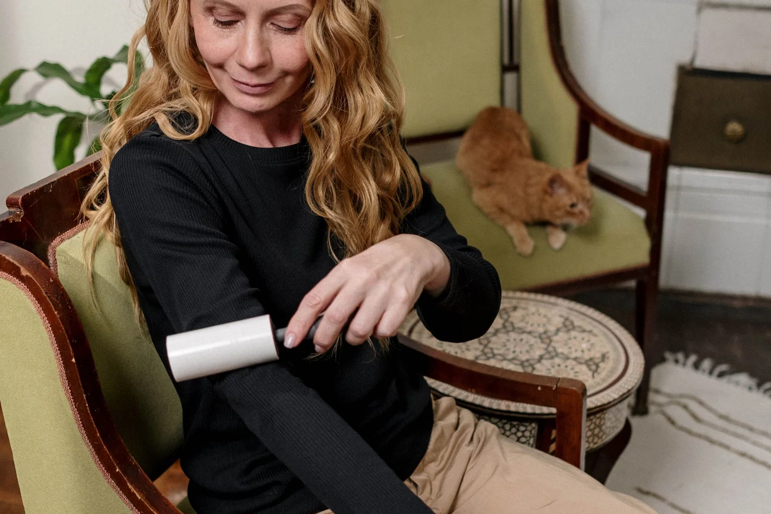 Woman using a lint roller on a black sweater while a ginger cat rests nearby